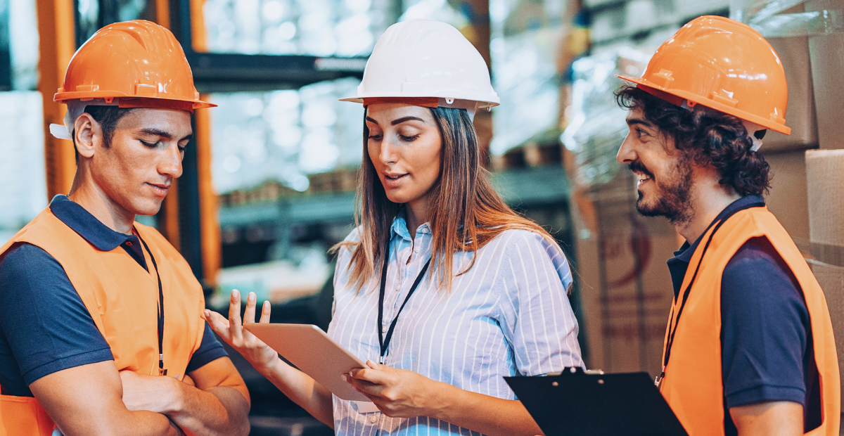 A female supervisor in a white helmet instructs two male workers in orange helmets in a warehouse.