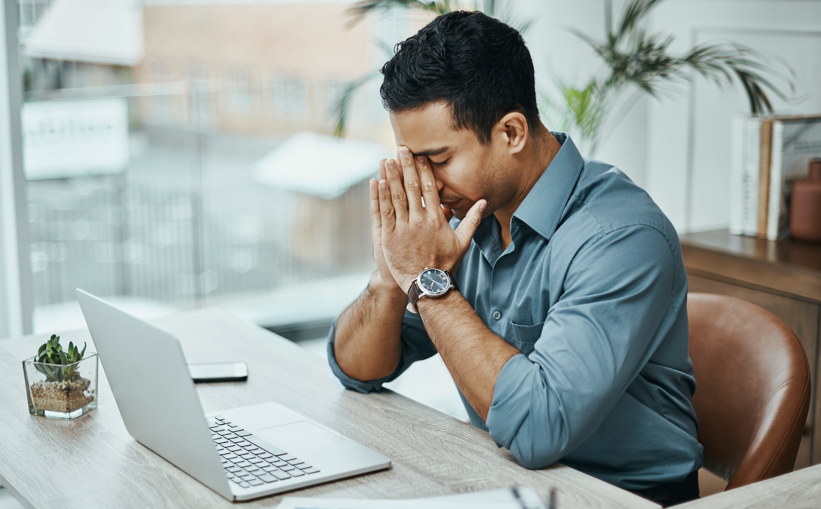 man sitting at his desk in front of his laptop looking stressed, with his head in his hands.