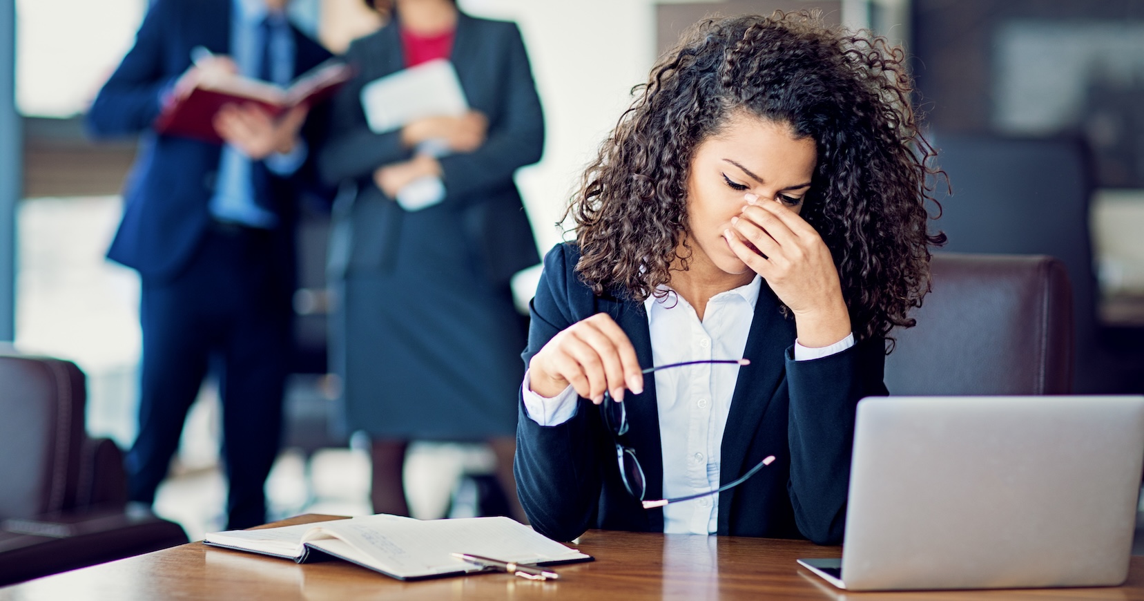 woman sitting in front of her computer looking stressed, with two colleagues standing behind her and looking like they are talking about her.